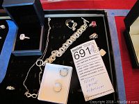 Photo showing a black box holding a silver ring with a clear stone, a silver chain necklace, circular link bracelet, and multiple rings and earrings on the black velvet background.