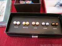Five pairs of spherical stud earrings in varying pearl-like colors with labels Peace, Believe, Love, Hope, Faith arranged in black jewelry box