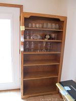 Full view of the five shelf wooden bookcase showing glassware on the upper shelves and empty lower shelves.
