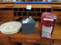 Photo showing red Keurig coffee maker, clear glass decanter on black box, cream-colored divided serving tray, and additional miscellaneous items on wooden desk.