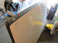 Large whiteboard with dark plastic frame and sticker label, angled leaning against furniture