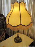 Front view of one table lamp showing brass-colored ornate base and beige scalloped lamp shade with fringe