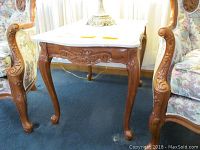 Side view of one side table showing light fruitwood carved base, cabriole legs with floral carving, and cultured white marble top with curved edges.