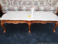 Front view of rectangular coffee table with white marble top and light fruitwood legs and apron showing carved details.