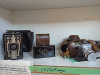 Shelf view showing three vintage cameras: Kodak Brownie, Tower 24, Nikon SLR with cases and straps