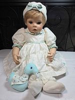 Front view of doll seated wearing dress, hat, booties, holding stuffed duck