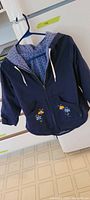 Front view of navy hooded children's jacket on hanger showing zipper, pockets, hood lining, embroidery