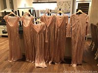 View of six rose gold sequin formal gowns hanging showing front details including sleeves, necklines, and length.