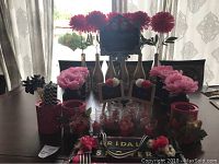 Photo shows decorated wine bottles, decorated mason jars, pink floral decorations, tea light holders, candles, and other components of a bridal shower set arranged on a table in front of window.