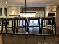 Four grey cloth bar stools with studded rims arranged in a row on a kitchen counter. Three stools are of equal height (26 inches) and one stool is shorter (24 inches). Minor scratches visible on wood frame. Dark wood legs with stainless steel trim and footrest bars.