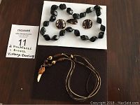 Lot of jewelry items on a dark wood surface: black beaded necklace, brown beaded necklace with pendant, fish brooch, and round black clip-on earrings.