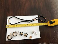 Black beaded necklace approximately 8 inches long with some orange beads near clasp, displayed together with four pairs of earrings on white background and measuring tape