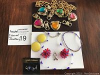 Assorted vintage coloured jewellery including chain belt with heart-shaped stones, clip-on earrings in yellow, red-purple cluster, and blue stone, purple bead necklace, and blue-yellow bracelet.