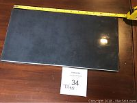 Top view of glossy black ceramic tile on wooden table with measuring tape showing approximately 7 x 14 inches size and a paper noting 34 tiles in the lot.