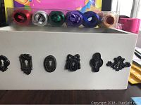 White wooden box with metal decorative keyhole covers and six colorful ribbon rolls displayed on top.