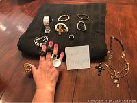 Photo showing assorted costume jewelry on table, including rings worn on hand, bracelets, necklaces, and a note with lot number
