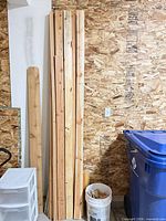 Stack of vertical pine boards leaning against OSB wall with bucket and recycling bin for scale