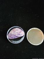Top view of charoite cabochon in plastic gem jar with lid beside