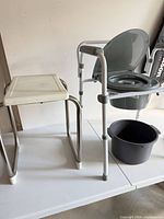 Bedside commode with seat flipped up, removable gray bucket, and bath stool side by side