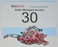 Photo of silver-tone metal brooch shaped like a leaf or feather with pink, clear, and green rhinestones, placed below a paper label with auction and lot number 30.