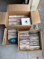 Two cardboard boxes and one liquor box filled with stacked 7-inch records in paper sleeves