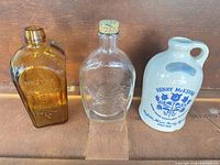 Front view of all three items: amber bottle, clear 1776 flask, stoneware jug