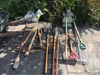Overall view of laid out garden tools on patio showing shovels, axes, pick axes, and various rakes.