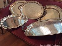 Five silver plate trays displayed on maroon upholstery showcasing different shapes and ornate handle designs