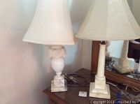Two alabaster base lamps side by side on a wooden surface, one with a finial and white fabric shade, the other with a cream fabric shade.