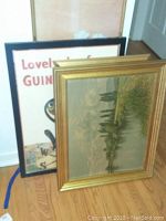 Three framed artworks stacked leaning against a wall, including the visible oil painting with landscape and a printed piece with partial text.