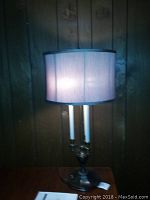 Front view of lit Stiffel Bouillotte candlestick table lamp against a wood-paneled wall, showing cream fabric drum shade and brass candlestick base with two candle-style bulbs.