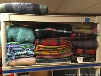Stack of assorted folded wool and mohair throws and lap blankets in various colors and patterns on a shelf.