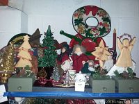 Photo of assorted Christmas decorations including ceramic trees, wooden angels, figurines, a wreath, stockings, and holiday tins arranged on green boxes against a concrete wall.