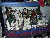 Full view of ceramic nativity scene figures with brass reindeer, ceramic Christmas tree, snowmen, Santa Claus and assorted holiday decorations arranged on a plaid cloth under a shelf.