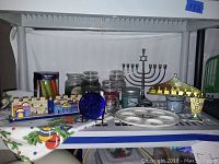 Wide view of Hanukkah items including menorahs, candles, seder plate, and decorative items arranged on a table with holiday-themed cloth.