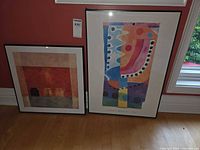 Both framed abstract prints leaning against wall on floor
