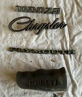 All four items laid out on cloth: Monza, Chrysler, Plymouth emblems and Hurst shift knob boot