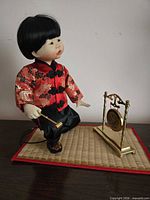 Front view of doll kneeling on bamboo mat holding mallet beside gong