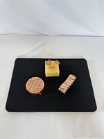 Bracelet, three rings on foam block, and closed compact mirror displayed on black surface