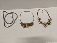 Three necklaces laid out: bendable silver-tone, gold-tone spike fringe, pastel floral statement