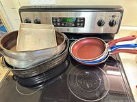All cookware stacked on stovetop: frying pans, oval roasters, square pan, speckled enamel roasters