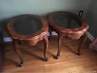 Two carved wood oval side tables with smoked glass tops placed next to a wall on hardwood floor.