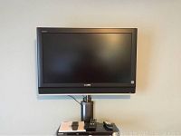 Full view of mounted Sony Bravia 32 inch TV with wall mount, remote controls and power cord visible on shelf below.