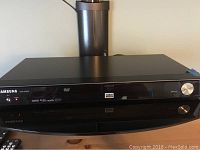 Front view of Samsung DVD-HD870C DVD player placed on black cylindrical stand with part of a remote visible.
