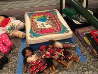 Closed Barbie case with colorful vintage Barbie illustration on lid, surrounded by a large baby doll and several folk dolls in traditional costumes