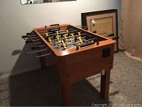 Foosball table angled view showing wood frame, metal rods, black handles, and player figures in yellow and black.