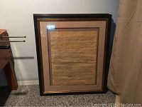 Front view of a framed page of The Constitution showing cursive script on parchment-style paper inside a dark wood frame with matte, standing on carpet against wall.