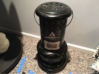 Front and top view of black metal vintage kerosene heater with metal handle and decorative perforations on top and sides.