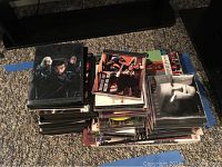 Pile of CDs and DVDs stacked on carpet beside TV, showing assorted media cases including a Bob Dylan LP partially visible beneath.