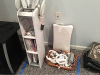 Wii accessories laid out on the floor including remotes, Nunchuks, steering wheels, power cables and balance board next to storage stand.